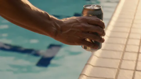 Two hands lifting a sweating glass of iced cold brew off a sunlit tiled ledge